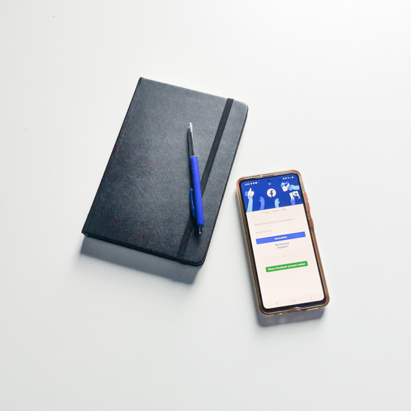 Zwart notitieboekje met balpen en iPhone met geopende Facebook-app op witte achtergrond. Illustratie voor blog over performance marketing en adverteren op Facebook.